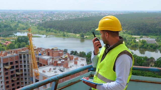 Portrait of Caucasian handsome man builder manager in uniform and helmet looking at digital tablet and talking in walkie-talkie while standing on construction site. Work, business