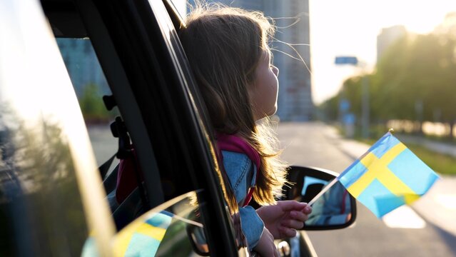 Pretty little girl in window with amazed face expression. Beautiful small Caucasian child looking out of car window holding in hand Swedish flag. Female teenager patriot. Close up - Powered by Adobe
