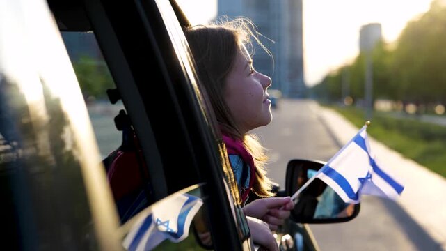 Joyful little girl holding Israeli flag in hand looking out of car window. Close up shot of small pretty child travels in Israel. Patriotic. Family trip. Travel concept. Slow motion
