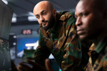 Fototapeta premium Team of military dispatchers looking at surveillance monitor, analyzing data to allocate resources to units based on mission needs. Agent and colleague examining strategic data on equipment displays