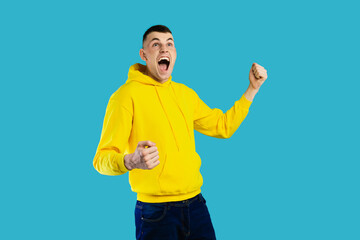A young man in a yellow hoodie expresses excitement while raising his fists in celebration. His...