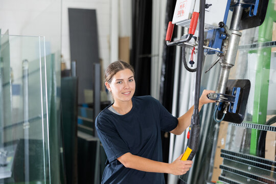 Experienced young female technician confidently smiling while operating vacuum lifting machine to move glass sheets in processing plant floor..