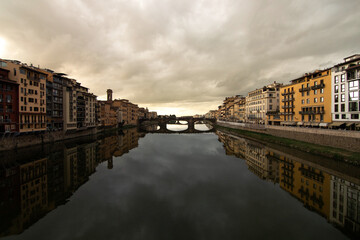 View of the Arno River in Florence, Tuscany, Italy, with a stone bridge and colorful historic buildings reflected in the calm water under a dramatic cloudy sky. Classic Italian cityscape.