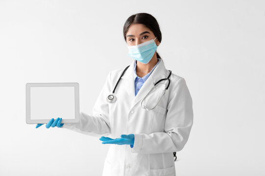 Young Hindu lady doctor in a white coat and stethoscope holds a tablet and gestures to a blank screen. She is wearing a protective mask and gloves while promoting remote health communication. - Powered by Adobe
