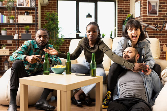 African American man and Caucasian woman compete in video game with wireless controllers while friends watch. Diverse group laughing, bonding and enjoying relaxed hangout in cozy living room. - Powered by Adobe