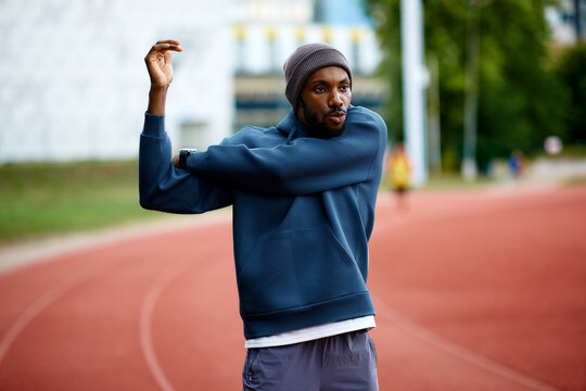 Focused black athlete stretches his triceps or shoulders as part of his warm-up routine on an outdoor running track before a workout