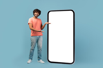 A stylish eastern man with curly hair is smiling and standing next to a big cell phone with a blank white screen. This scene takes place in a blue studio.
