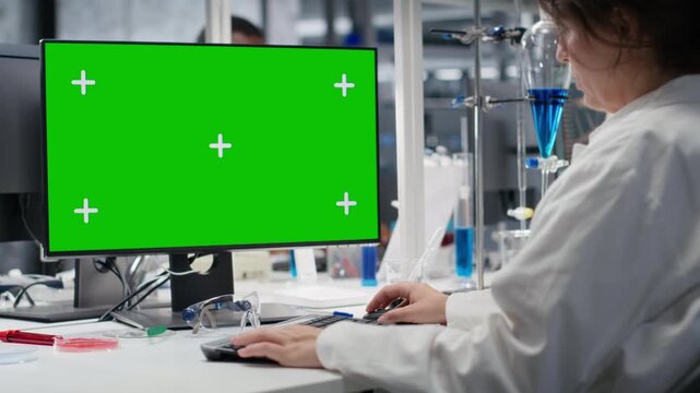 Lab technician at desk typing on green screen PC keyboard, looking at molecular model, doing research tasks. Woman using bioengineering program on mockup computer in medical lab, camera B