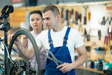 Young male repair service worker in uniform fixing female customer's bike