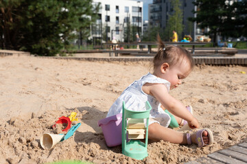 a young child in a white dress plays in an urban sandbox on a sunny day, surrounded by colorful toys, with buildings and trees in the background, creating a peaceful city playground scene, setting