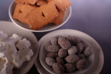 close-up of assorted treats featuring powdered chocolate almonds, sugar cubes, and decorative biscuits on plates with a soft purple background for a delicious display, artistic, savory, nibble