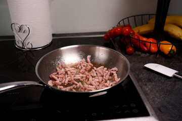 cooking bacon bits in a frying pan on an induction stove with fresh vegetables and a roll of paper towels adding color and utility to the modern kitchen setting, surface, heat, slices, seasoning