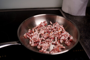 close-up of diced bacon frying in a pan on a dark stove top in modern kitchen setting with paper towel roll and subtle lighting reflecting on stainless steel surface, unattended, breakfast, strip