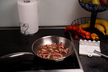 kitchen scene with bacon frying in pan on stove near paper towel and fruit basket containing tomatoes, bananas, and peppers with spatula resting, gourmet, lifestyle, decor, appetizing, modern, life