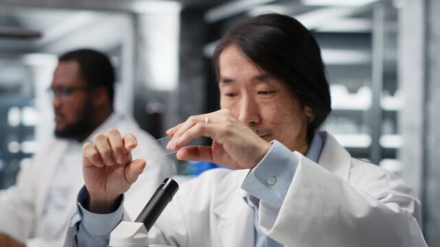 Clinical microbiologist examining microscope glass slide in laboratory to view cells. Asian lab worker checks sample slide before imaging process using optical device, camera B