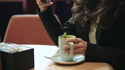 Caucasian female sitting inside of modern cafe with a lot of green plants, drinking hot tea with mint with pleasure, smiling and scrolling her smartphone.