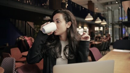 Caucasian woman sitting inside of cafe. Girl is looking around, smiling, and drinking hot coffee from a white cup. Female is working on her computer.