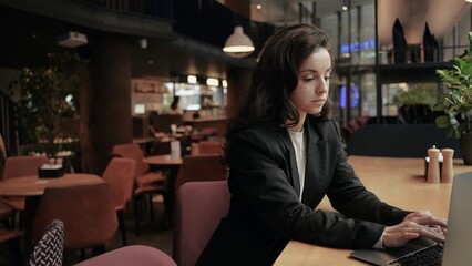 Young Caucasian woman sitting in the cafe at a wooden table, girl is typing on her laptop and she looks very tired.