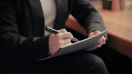 Young beautiful Caucasian woman in black jacket and white shirt sitting inside the cafe and drawing in her notebook.