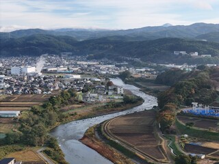 A river runs through a town with houses and buildings on both sides. The river is surrounded by fields and mountains in the background