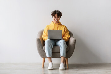 Young Asian man enjoys working on his laptop while sitting comfortably in an armchair against a neutral studio wall. He appears engaged and joyful as he browses the internet.