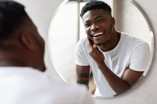 Man stands in modern bathroom, touching his chin with bristle before shaving. He smiles at his reflection, enjoying his skincare routine in a relaxed setting.