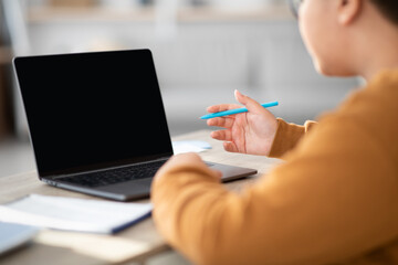 Unrecognizable chubby boy with glasses sits at a table using a notebook and computer. He appears focused while doing homework and having a video call with his teacher in a bright room.