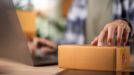 A small business owner prepares a cardboard box for delivery while managing online orders on a laptop, symbolizing e-commerce, logistics, and digital retail efficiency.