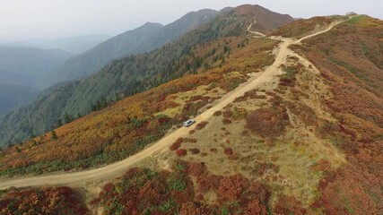 Aerial view of 4x4 SUV driving on a dirt mountain road along a hill ridge. Off-road vehicle traveling through autumn forest and brushland in fall season. - Powered by Adobe