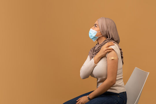 Coronavirus vaccination banner. Arab lady in face mask having adhesive bandage on shoulder after covid vaccine shot, looking at free space on brown background, side view