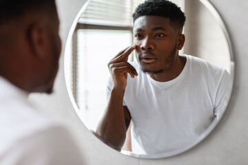 In a bright bathroom, a handsome black man applies moisturizer under his eyes while gazing into a...