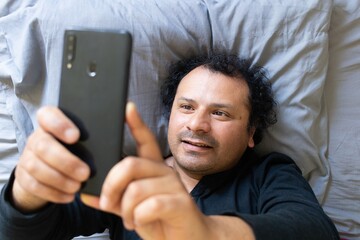 Latino man lying on bed checks his cell phone
