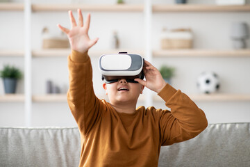A chubby Korean teen boy is fully immersed in a virtual reality session at home. He is excitedly reaching out to touch something invisible while wearing modern VR glasses.