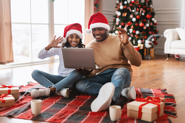 A happy black dad and his child wearing Santa hats sit on the floor, waving during a video call. They celebrate Christmas together, surrounded by gifts and a decorated tree.