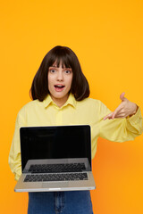 Naklejka premium A surprised woman stands against a vivid yellow background, presenting a laptop with both hands, creating a playful and energetic moment of online exploration.