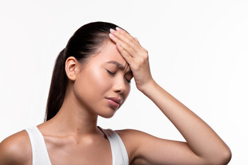 Fototapeta premium A young Chinese woman appears unhappy as she touches her forehead with closed eyes. She is suffering from a headache in a bright white studio setting with ample copy space.