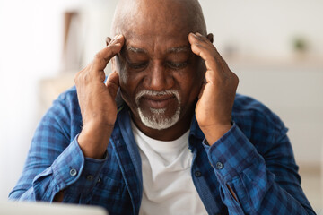 Senior African American man sits indoors, massaging his temples to relieve a headache. He appears distressed, emphasizing health concerns faced by older adults in their daily lives.