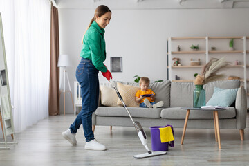 A woman in a green shirt and gloves mops the floor in a bright living room while a young child sits on the couch, focused on a tablet. The space is stylish and airy.