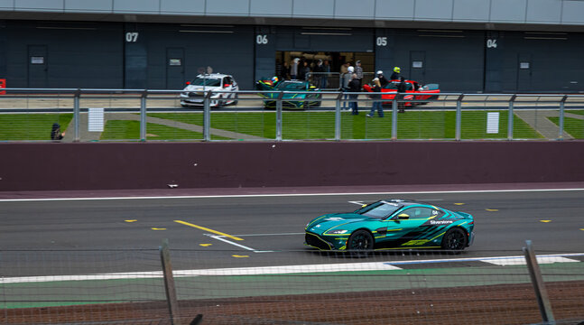 Sports car driving past the pit lane at Silverstone Circuit, with crews and support vehicles in the background.
