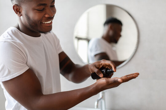 A happy black man stands in a modern bathroom, squeezing a bottle of shaving cream into his hand. He focuses on his facial care routine while looking in the mirror. - Powered by Adobe