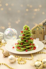 Christmas tree made of kiwi, pomegranate seeds and festive decor on beige table against light background with blurred lights, closeup. Bokeh effect