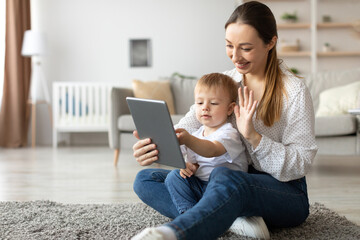 A joyful mother sits on the floor with her young son, using a tablet to video chat. They both smile and wave, enjoying their time together in a bright and inviting living room.