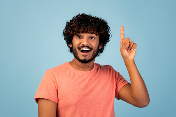 A joyful Hindu guy in a pink t-shirt stands in a blue studio. He is smiling broadly and pointing his index finger up, showcasing excitement about a recent idea. © Prostock-studio