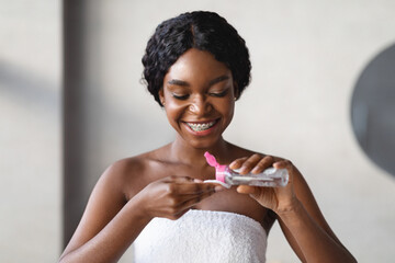 A pretty young black woman smiles as she applies micellar water onto a cotton pad. She is preparing her face for makeup while wrapped in a white towel in her bathroom.