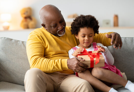 In a warm living room, a happy African American preschool kid sits beside an older man, both excitedly opening a birthday present together. Their joy captures a loving family moment.