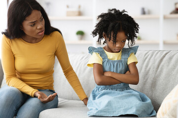 A mother talks to her daughter on a sofa in a living room. The child appears upset and is sitting with her arms crossed. The setting is calm and cozy.