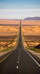 Fototapeta premium A long empty asphalt road stretches through a vast desert landscape with sand dunes and mesas under a clear blue sky