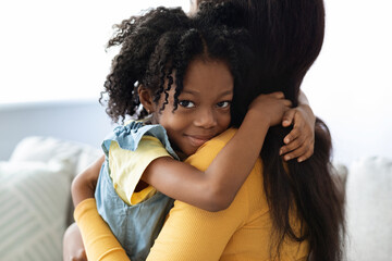 Obraz premium A young girl hugs her mother tightly, smiling sweetly. They are in a bright living room with soft furniture, enjoying a tender moment together.