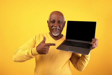 Elderly black man stands against an orange background, smiling and pointing at a laptop with a...