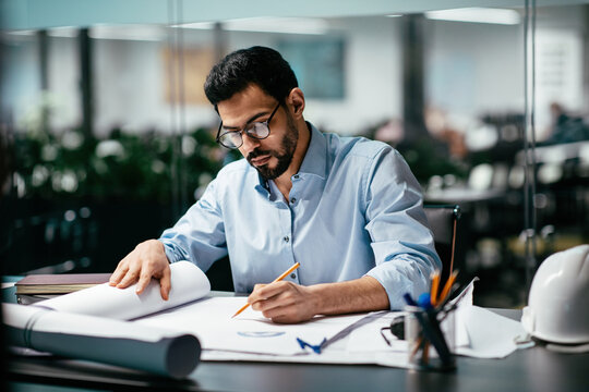 A focused young man designs architectural plans at a contemporary office, surrounded by office plants and tools. He meticulously sketches with a pencil on large sheets.